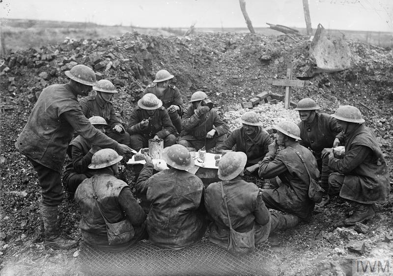 British soldiers eating Christmas dinner on a makeshift table in a shell crater. A wooden grave marker can be seen in the background.